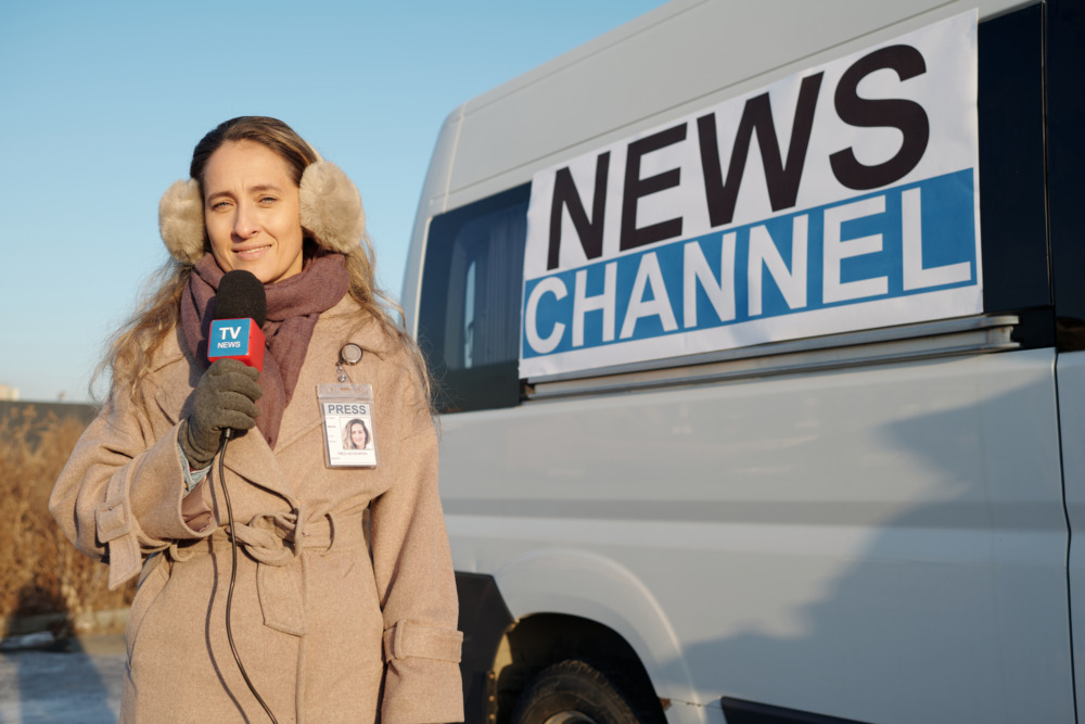 Happy young female journalist looking at camera and speaking in microphone against TV van from news channel