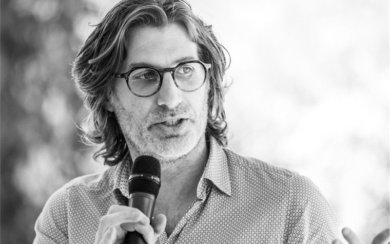 PromptZero's lead Christopher Moscardi, holding a microphone and speaking at an event, wearing a patterned button-up shirt, in a black and white photograph, outdoor background.