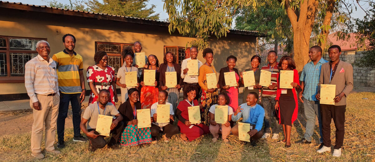 A group of teachers proudly displaying certificates while standing and kneeling together outdoors in front of the TTCZS Resoruce Centre and a tree during sunset.