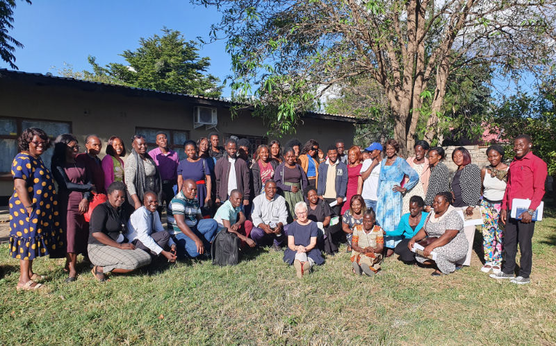 A large group of adults posing together outdoors in front of TTCSZ's Resource Centre and a tree on a sunny day, with some people standing and others kneeling or sitting on the grass.