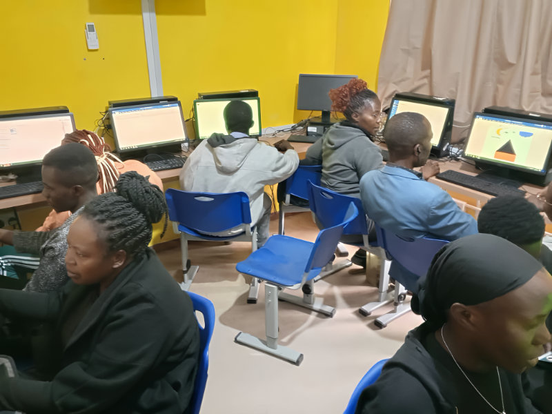 A group of people working on desktop computers in a bright classroom with yellow walls. The room features blue chairs and multiple computers lined up on desks along the wall. Some computer screens display geometric shapes, while others appear to show text documents. The classroom setting suggests a computer training or educational session.