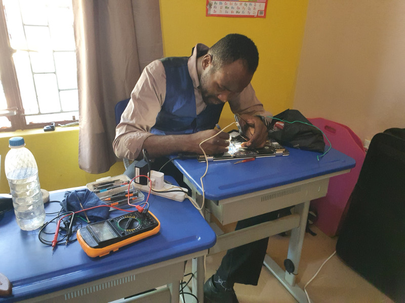 Kunda Phanuel, TTCSZ's chief repairer. He is seated at a blue desk in a classroom, focused on repairing an open laptop using electronic tools. The desk is covered with various equipment, including a digital multimeter, wires, and a water bottle. Bright sunlight enters through a nearby window, and educational posters are visible on the yellow classroom wall.