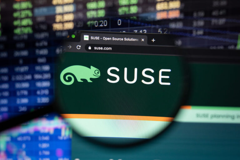 SUSE Linux SLES 16 shown by Suse company logo on a website with blurry stock market developments in the background, seen on a computer screen through a magnifying glass