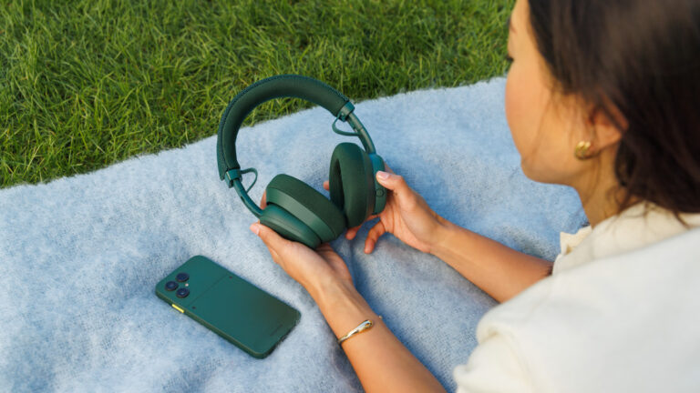 Person relaxing outdoors on a blanket, holding green Fairphone Fairbuds XL over-ear headphones next to a matching green Fairphone smartphone on the grass.