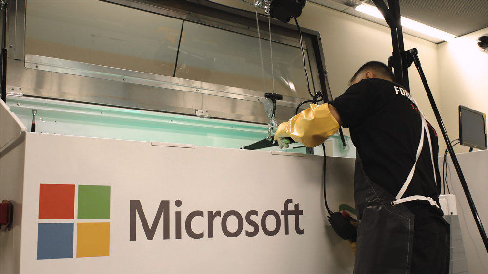 A technician wearing protective gloves and an apron works at a large white Microsoft-branded liquid cooling tank in a data center.