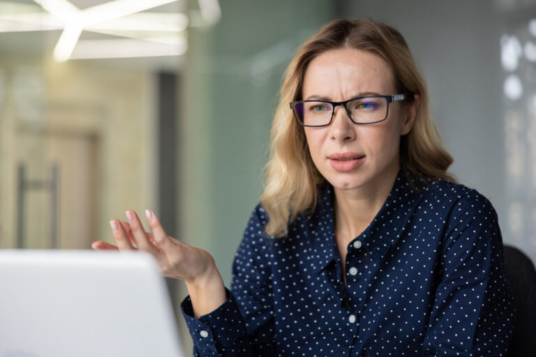 AI trust gap shown by young businesswoman with glasses experiencing confusion and frustration while having a video call on a laptop, gesturing during a remote meeting or online conversation in an office setting