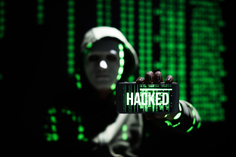 Russian hacker holding mobile phone with word HACKED on screen against dark background