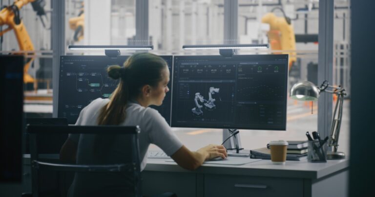 Schneider’s TeSys Tera shown via pic of Focused Female Engineer Tracking Robotic Performance Metrics and Digital Twin Model on Workstation, With Factory in Background. Data Driven Decision Making and Real Time System Analysis. Back View.