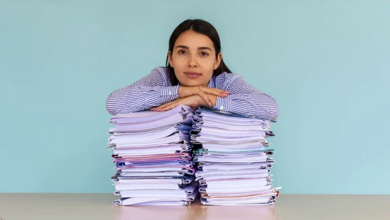 digital ERP agents Reducing administration burden shown by a tired businesswoman leaning her arms and chin on two high stacks of paper folders on a desk against a plain blue background, depicting office exhaustion, heavy workload, and corporate stress