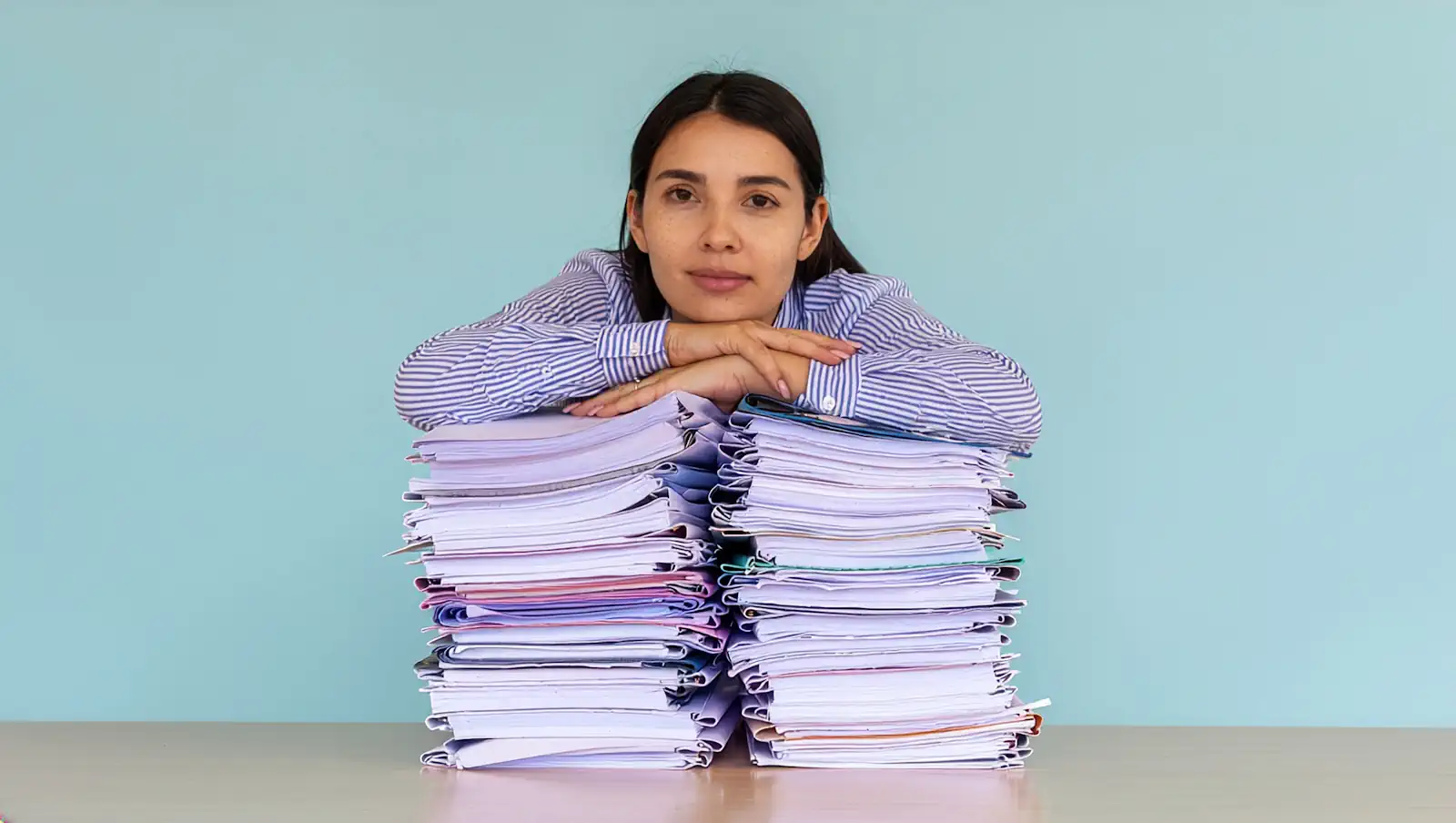 digital ERP agents Reducing administration burden shown by a tired businesswoman leaning her arms and chin on two high stacks of paper folders on a desk against a plain blue background, depicting office exhaustion, heavy workload, and corporate stress