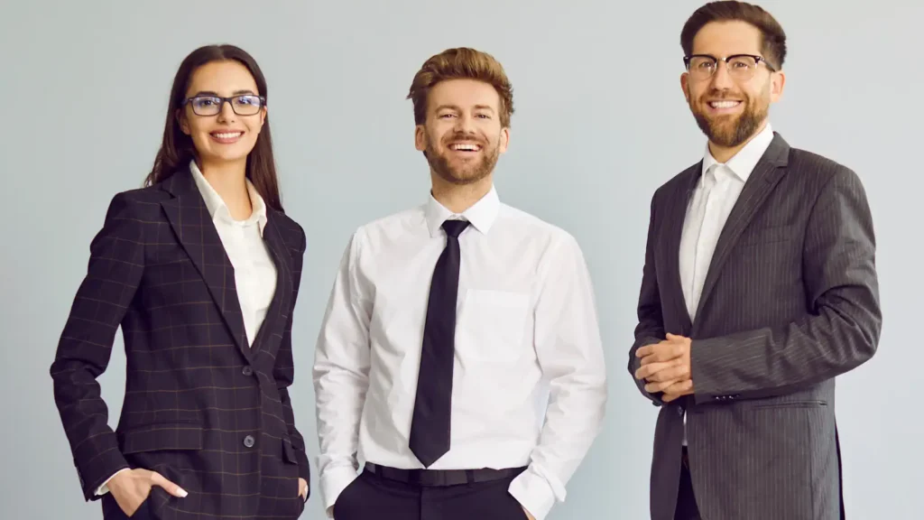 a group of three young cheerful business people in suits standing confidently, looking at the camera and smiling isolated on a studio grey background