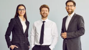 a group of three young cheerful business people in suits standing confidently, looking at the camera and smiling isolated on a studio grey background