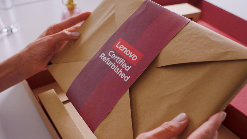 Close-up of a person's hands holding a laptop wrapped in brown paper, secured with a red and burgundy band that reads "Lenovo Certified Refurbished", resting inside a red cardboard box.