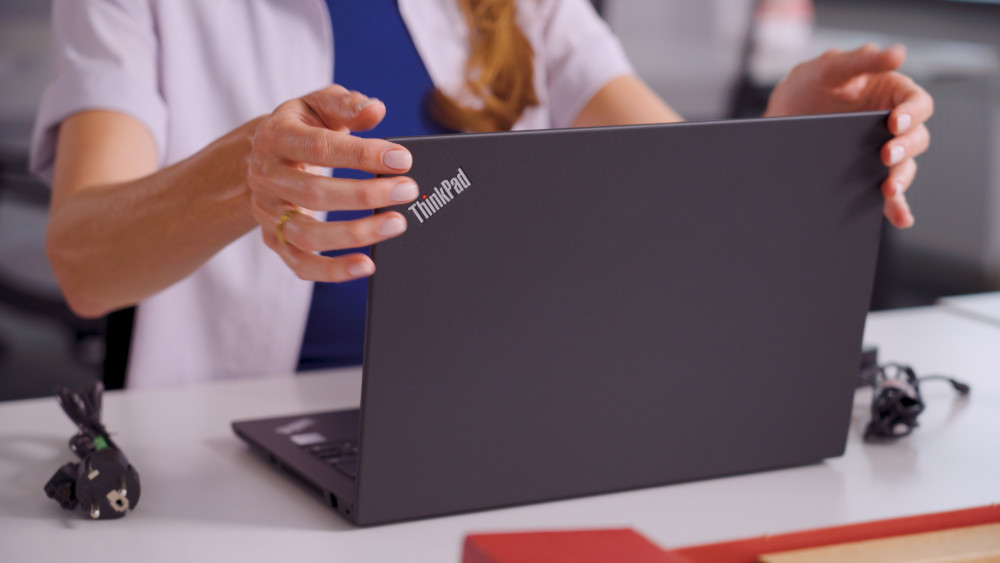 A person opening the lid of a black Lenovo ThinkPad laptop on a white desk, with a power cable and adapter resting alongside the computer