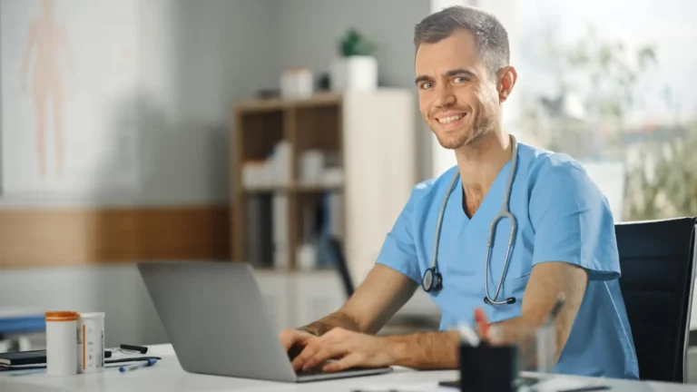Male Nurse Wearing Blue Uniform Working on Laptop Computer at Doctor's Office and Smiling on Camera