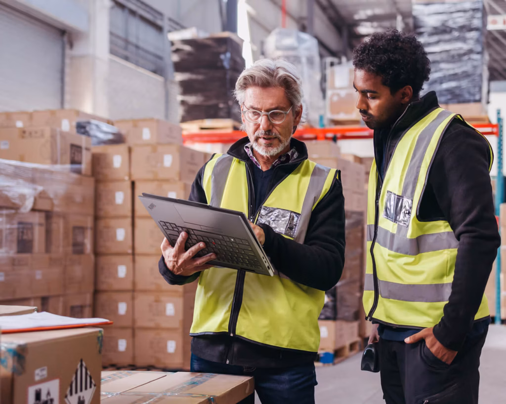 An older man with gray hair, glasses, and a beard, and a younger Black man, both wearing high-visibility vests over dark jackets, look closely at a Lenovo ThinkPad laptop held by the older man in a busy warehouse. The background is filled with stacks of cardboard boxes on pallets and warehouse shelving. The older man is gesturing toward the screen as they discuss, while the younger man, with his hand in his pocket and holding a scanner, looks on attentively. In the foreground, more taped cardboard boxes are visible, capturing an active logistics or inventory environment.
